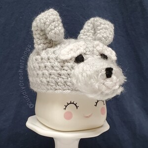 May include: A handmade, light gray crochet hat shaped like a dog's head, complete with ears, a fluffy muzzle, and black eyes and nose. The hat is placed on top of a white mug with a smiling face. The text "cuddlyCrochetThings" is visible.