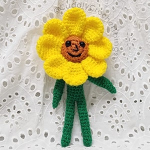 May include: A crocheted sunflower with a smiling face, yellow petals, and green stem and leaves. The sunflower is standing on a white background.