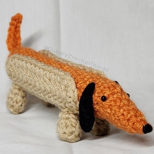 May include: A crocheted dachshund toy with a tan body and orange head. The toy has black eyes and a black nose.