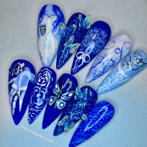 Puede incluir: Un conjunto de diez uñas artificiales largas y puntiagudas en varios tonos de azul. Cada uña presenta diseños únicos, incluyendo mariposas, patrones florales y arte abstracto. Algunas uñas tienen adornos de mariposas en 3D. El texto "helenmiladin.nails" es visible.