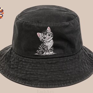 May include: A dark grey bucket hat featuring an embroidered kitten design. The kitten is grey and white with pink ears. The hat is made of a soft, textured material, suitable for casual wear.