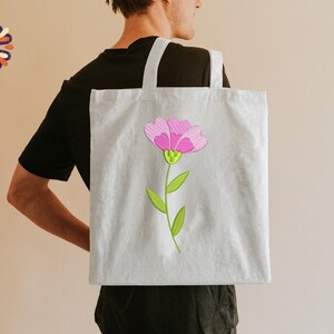 May include: A light gray tote bag featuring a pink flower with green leaves and stem. The flower has a detailed design with pink petals and a yellow center. The bag has white handles and is carried over a person's shoulder.