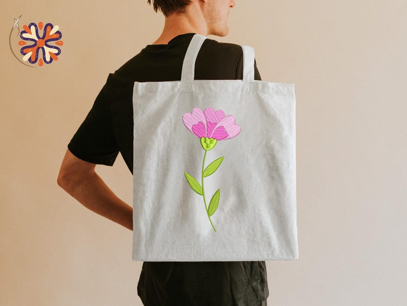 May include: A light gray tote bag featuring a pink flower with green leaves and stem. The flower has a detailed design with pink petals and a yellow center. The bag has white handles and is carried over a person's shoulder.