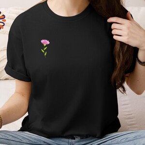 May include: Black t-shirt with a small pink flower design on the chest. The flower has a green stem and leaves. The shirt is a solid black color and has short sleeves.
