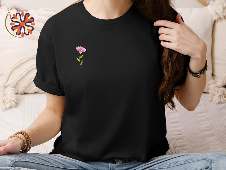 May include: Black t-shirt with a small pink flower design on the chest. The flower has a green stem and leaves. The shirt is a solid black color and has short sleeves.