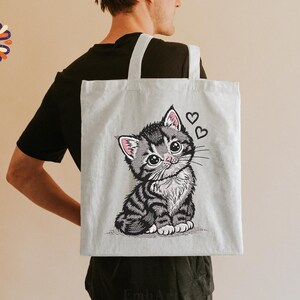 May include: A light grey tote bag with an embroidered illustration of a cute kitten with big eyes and two hearts. The kitten has grey and black stripes and pink accents on its ears. The tote bag has white handles.