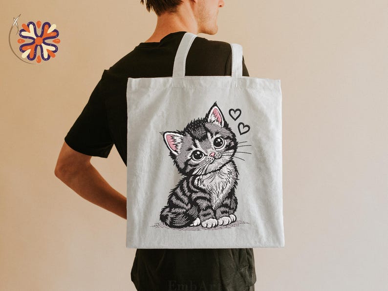 May include: A light grey tote bag with an embroidered illustration of a cute kitten with big eyes and two hearts. The kitten has grey and black stripes and pink accents on its ears. The tote bag has white handles.