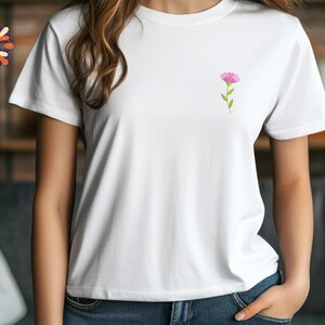 May include: White t-shirt with a small pink flower design on the chest. The flower has a green stem and leaves. The shirt is short-sleeved and made of a soft material. The model is wearing blue jeans.