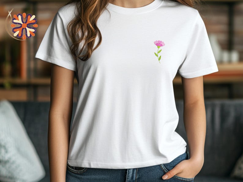 May include: White t-shirt with a small pink flower design on the chest. The flower has a green stem and leaves. The shirt is short-sleeved and made of a soft material. The model is wearing blue jeans.