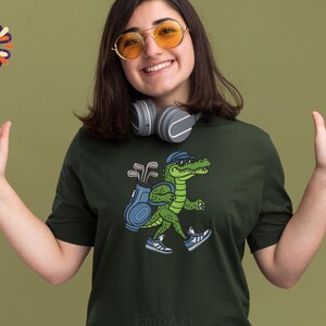 May include: Dark green t-shirt featuring a cartoon alligator wearing a cap, carrying a golf bag, and wearing sneakers. The alligator is walking. The background is a solid olive green color. The person in the image is wearing headphones and sunglasses.
