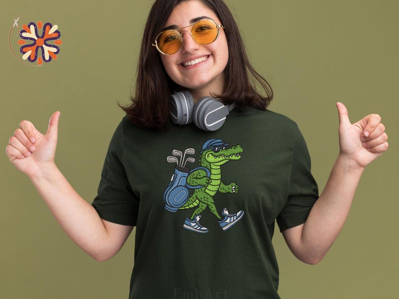 May include: Dark green t-shirt featuring a cartoon alligator wearing a cap, carrying a golf bag, and wearing sneakers. The alligator is walking. The background is a solid olive green color. The person in the image is wearing headphones and sunglasses.