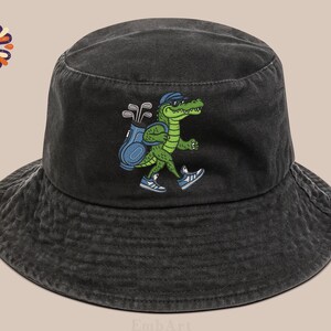 May include: Black bucket hat with a green alligator graphic wearing sunglasses and carrying a golf bag. The alligator is wearing sneakers. The hat is made of a washed, textured fabric.