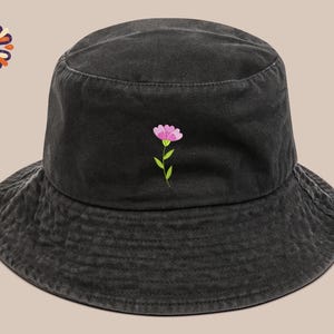 May include: Black bucket hat with a pink flower embroidery. The hat features a wide brim and a simple, casual design. The flower has pink petals and a green stem and leaves. The hat is made of a soft, textured fabric.
