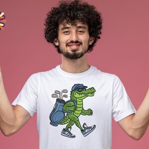 May include: White t-shirt featuring a cartoon alligator wearing sunglasses and a golf hat, carrying a golf bag and clubs. The alligator is walking and wearing blue sneakers. The background is a solid pink color.