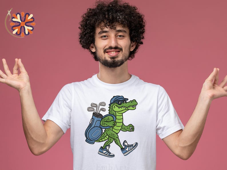 May include: White t-shirt featuring a cartoon alligator wearing sunglasses and a golf hat, carrying a golf bag and clubs. The alligator is walking and wearing blue sneakers. The background is a solid pink color.