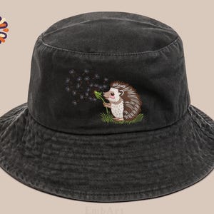 Puede incluir: Sombrero de pescador de mezclilla negra con un erizo bordado soplando semillas de diente de le&oacute;n. El erizo est&aacute; representado en un estilo de dibujos animados con detalles marrones, blancos y rosas. El sombrero tiene un ala ancha y un aspecto descolorido.