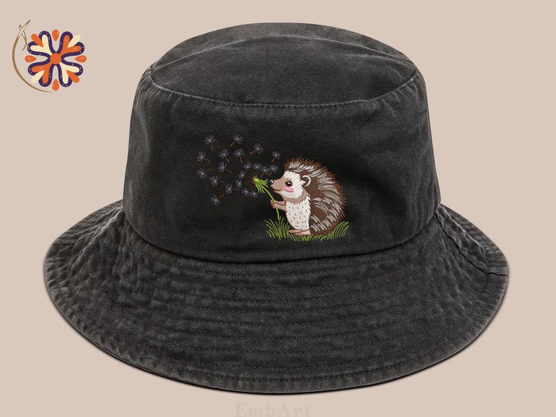 Puede incluir: Sombrero de pescador de mezclilla negra con un erizo bordado soplando semillas de diente de le&oacute;n. El erizo est&aacute; representado en un estilo de dibujos animados con detalles marrones, blancos y rosas. El sombrero tiene un ala ancha y un aspecto descolorido.