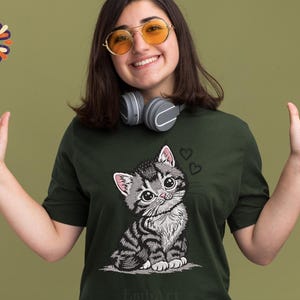 May include: Dark green t-shirt with a cartoon kitten design. The kitten has large eyes and a striped pattern, with two hearts above its head. The t-shirt is worn by a person wearing sunglasses and headphones.