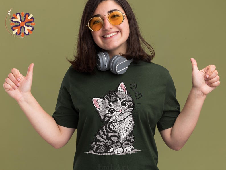 May include: Dark green t-shirt with a cartoon kitten design. The kitten has large eyes and a striped pattern, with two hearts above its head. The t-shirt is worn by a person wearing sunglasses and headphones.