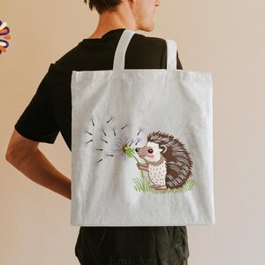 Puede incluir: Un bolso tote de color claro con una ilustraci&oacute;n bordada de un erizo soplando semillas de diente de le&oacute;n. El erizo tiene pelaje marr&oacute;n y blanco y sostiene un diente de le&oacute;n verde. El bolso tiene dos asas blancas.