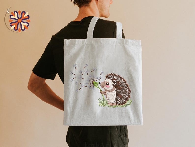 Puede incluir: Un bolso tote de color claro con una ilustraci&oacute;n bordada de un erizo soplando semillas de diente de le&oacute;n. El erizo tiene pelaje marr&oacute;n y blanco y sostiene un diente de le&oacute;n verde. El bolso tiene dos asas blancas.