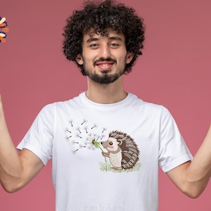 Puede incluir: Camiseta blanca con un erizo de dibujos animados soplando semillas de diente de le&oacute;n. El erizo es marr&oacute;n y beige, sosteniendo un diente de le&oacute;n. El dise&ntilde;o est&aacute; en una camiseta blanca. El fondo es de color rosa liso.