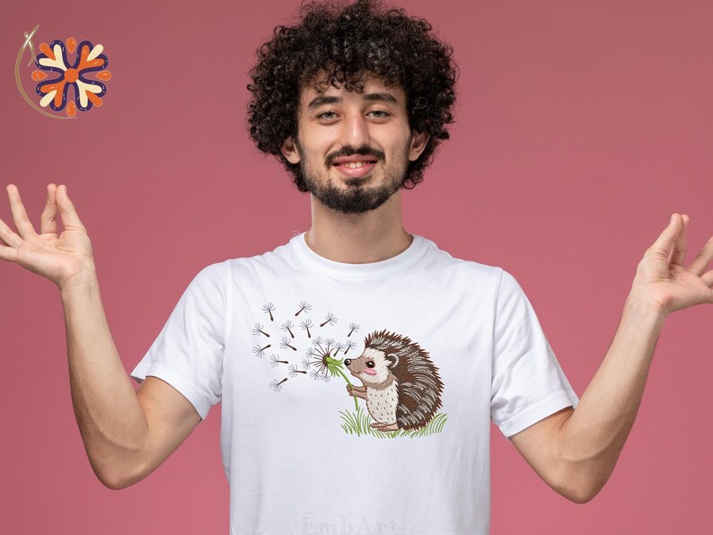 Puede incluir: Camiseta blanca con un erizo de dibujos animados soplando semillas de diente de le&oacute;n. El erizo es marr&oacute;n y beige, sosteniendo un diente de le&oacute;n. El dise&ntilde;o est&aacute; en una camiseta blanca. El fondo es de color rosa liso.