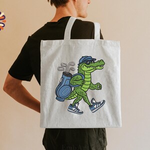 May include: A light gray tote bag featuring a cartoon alligator wearing sunglasses and a golf cap, carrying a golf bag with clubs. The alligator is wearing blue and white sneakers. The bag has white straps.