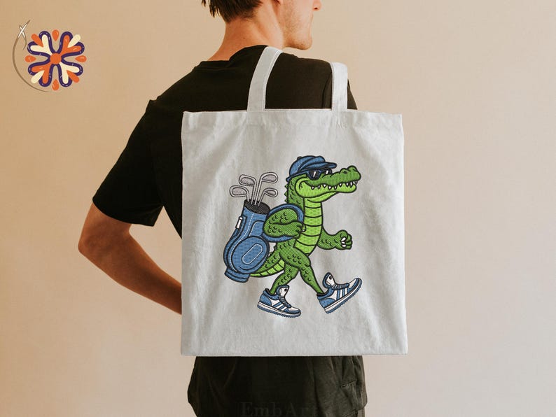 May include: A light gray tote bag featuring a cartoon alligator wearing sunglasses and a golf cap, carrying a golf bag with clubs. The alligator is wearing blue and white sneakers. The bag has white straps.