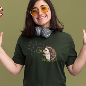Puede incluir: Camiseta verde oscuro con un bordado de un erizo soplando semillas de diente de le&oacute;n. El erizo tiene p&uacute;as marrones y blancas y una nariz rosa. La camiseta es de cuello redondo.