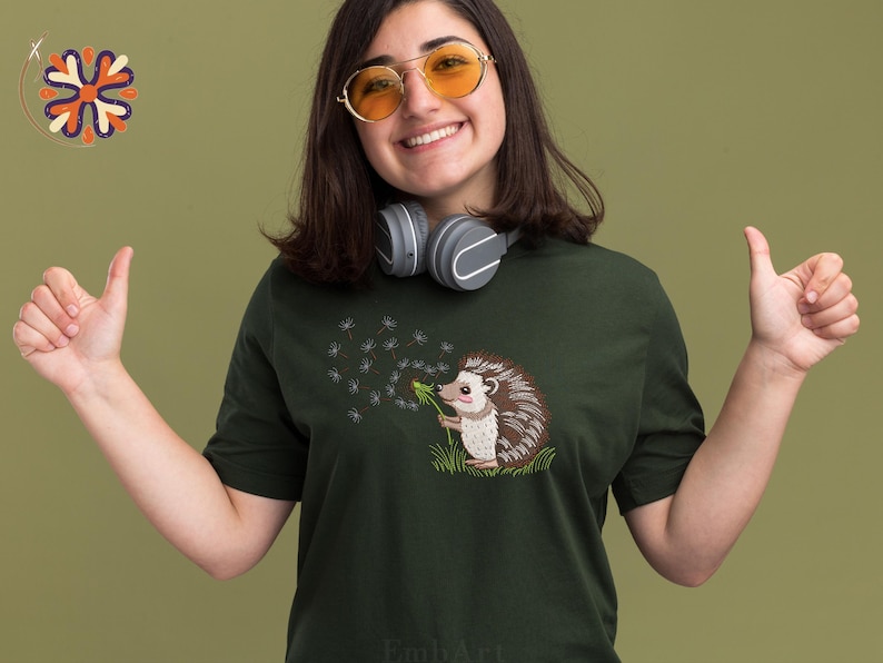 Puede incluir: Camiseta verde oscuro con un bordado de un erizo soplando semillas de diente de le&oacute;n. El erizo tiene p&uacute;as marrones y blancas y una nariz rosa. La camiseta es de cuello redondo.