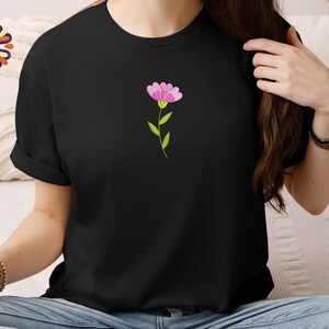 May include: Black t-shirt featuring a small, embroidered pink flower with green stem and leaves. The flower is centered on the chest. The shirt has short sleeves and a crew neck. The overall style is casual and simple.