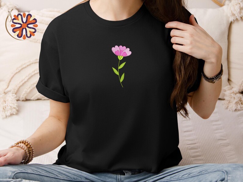 May include: Black t-shirt featuring a small, embroidered pink flower with green stem and leaves. The flower is centered on the chest. The shirt has short sleeves and a crew neck. The overall style is casual and simple.