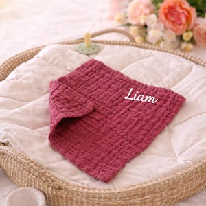 May include: A burgundy-colored baby bib with the name "Liam" embroidered in white. The bib is made of a textured fabric and is folded on a white quilted surface inside a woven basket. A pacifier is visible in the background.
