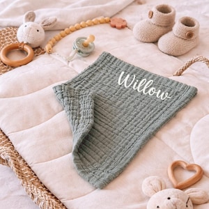 May include: A sage green baby blanket with the name "Willow" in white script. The blanket is surrounded by baby items, including knitted booties, a pacifier, a wooden teether, and a plush bunny toy.