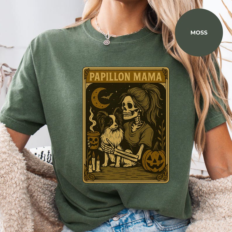 Puede incluir: Camiseta verde musgo con un dise&ntilde;o gr&aacute;fico que muestra un esqueleto sosteniendo un perro Papillon, una taza de caf&eacute; y una calabaza. El texto "Papillon Mama" est&aacute; en la parte superior. La camiseta es de color s&oacute;lido.