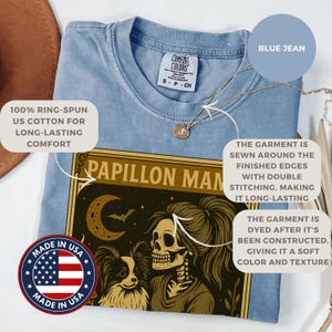 Puede incluir: Camiseta azul claro con un gr&aacute;fico que presenta una mujer esqueleto y un perro, con el texto "PAPILLON MAN". La camiseta est&aacute; hecha de algod&oacute;n estadounidense 100% hilado en anillo y te&ntilde;ida para un color y textura suaves.