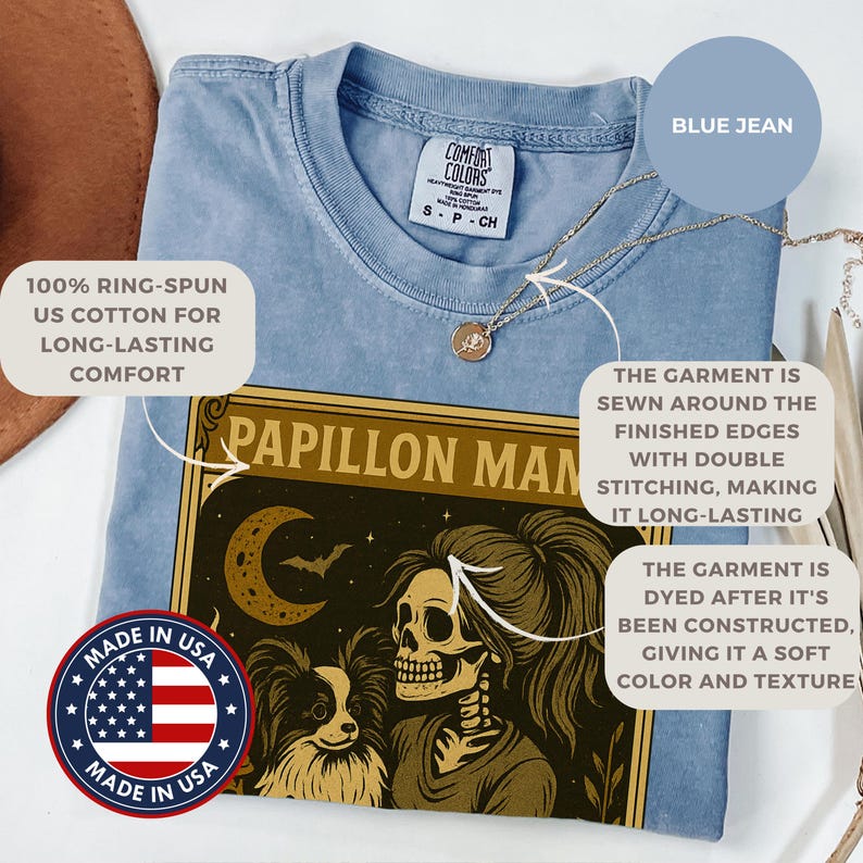 Puede incluir: Camiseta azul claro con un gr&aacute;fico que presenta una mujer esqueleto y un perro, con el texto "PAPILLON MAN". La camiseta est&aacute; hecha de algod&oacute;n estadounidense 100% hilado en anillo y te&ntilde;ida para un color y textura suaves.