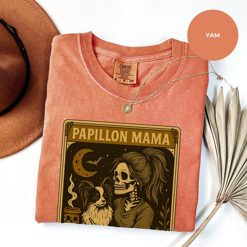 Puede incluir: Una camiseta color &ntilde;ame con las palabras "Papillon Mama" en un marco decorativo. El gr&aacute;fico presenta un esqueleto con un perro Papillon, una luna creciente y una taza de caf&eacute;. Un collar dorado est&aacute; drapeado sobre la camisa.
