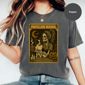Puede incluir: Camiseta gris con un dise&ntilde;o tem&aacute;tico de Halloween. El gr&aacute;fico muestra un esqueleto con un perro Papillon, una calabaza y el texto "Papillon Mama". El dise&ntilde;o utiliza tonos marrones y dorados.