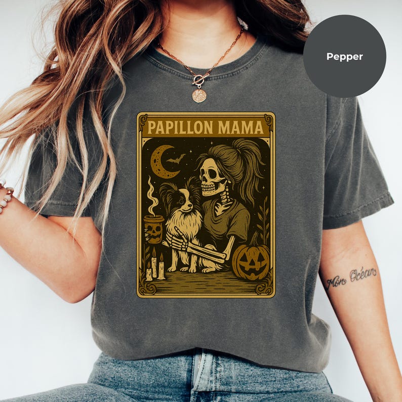 Puede incluir: Camiseta gris con un dise&ntilde;o tem&aacute;tico de Halloween. El gr&aacute;fico muestra un esqueleto con un perro Papillon, una calabaza y el texto "Papillon Mama". El dise&ntilde;o utiliza tonos marrones y dorados.