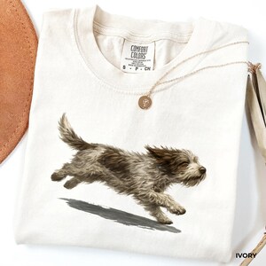 May include: An ivory-colored t-shirt featuring a graphic of a running brown and white scruffy dog. The shirt has a crew neckline and a Comfort Colors brand label at the inner neck. A thin gold-toned chain necklace with a small round pendant rests on top.