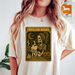 Puede incluir: Camiseta color crema con un dise&ntilde;o tem&aacute;tico de Halloween. El dise&ntilde;o incluye el texto "Papillon Mama", una mujer esqueleto con un perro Papillon, una luna creciente, una calabaza y velas. El dise&ntilde;o es en tonos marrones y dorados.