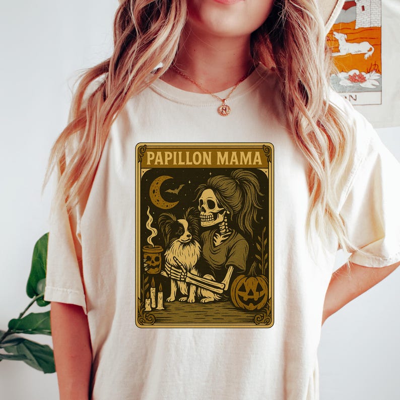 Puede incluir: Camiseta color crema con un dise&ntilde;o tem&aacute;tico de Halloween. El dise&ntilde;o incluye el texto "Papillon Mama", una mujer esqueleto con un perro Papillon, una luna creciente, una calabaza y velas. El dise&ntilde;o es en tonos marrones y dorados.