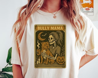 Könnte beinhalten: Ein cremefarbenes T-Shirt mit einem Grafikdruck, der ein Skelett zeigt, das einen Hund umarmt. Das Design enthält den Text BULLY MAMA, eine Mondsichel, eine Fledermaus, eine dampfende Tasse, zwei Kerzen und einen Kürbis in Braun- und Goldtönen.
