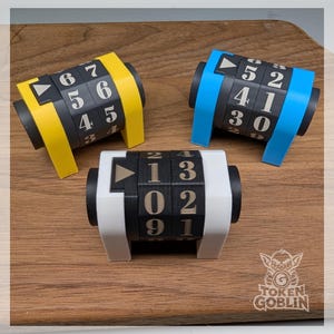 May include: Three mechanical life counters with rotating black number wheels and white numerals. Each counter features a coloured frame in yellow, blue, or white, with a small triangular indicator pointing towards the current number display.