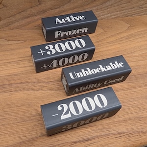May include: Four rectangular dark gray blocks with white text. The blocks feature various labels including Active, Frozen, +3000, +4000, Unblockable, Ability Used, -2000, and -3000. Each block has a small decorative emblem on the side edge.