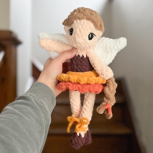 May include: A handmade crochet doll with a light skin tone, brown hair, and large black eyes. The doll wears a layered skirt in shades of orange and brown, with white wings and yellow bows on its feet. The doll is approximately 8 inches tall.