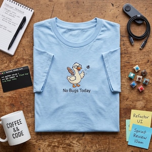 May include: A light blue t-shirt featuring a cartoon duck wearing glasses, holding a feather quill, and swatting away a small bug. Below the duck, the text reads "No Bugs Today" in a simple black font. The shirt is displayed on a wooden surface.