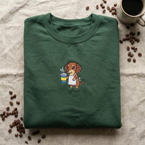 Peut inclure: Un sweat-shirt vert foncé à col rond. Le sweat-shirt présente un dessin brodé d'un teckel de dessin animé portant un tablier et tenant une tasse de café. Des grains de café et une tasse de café sont en arrière-plan.